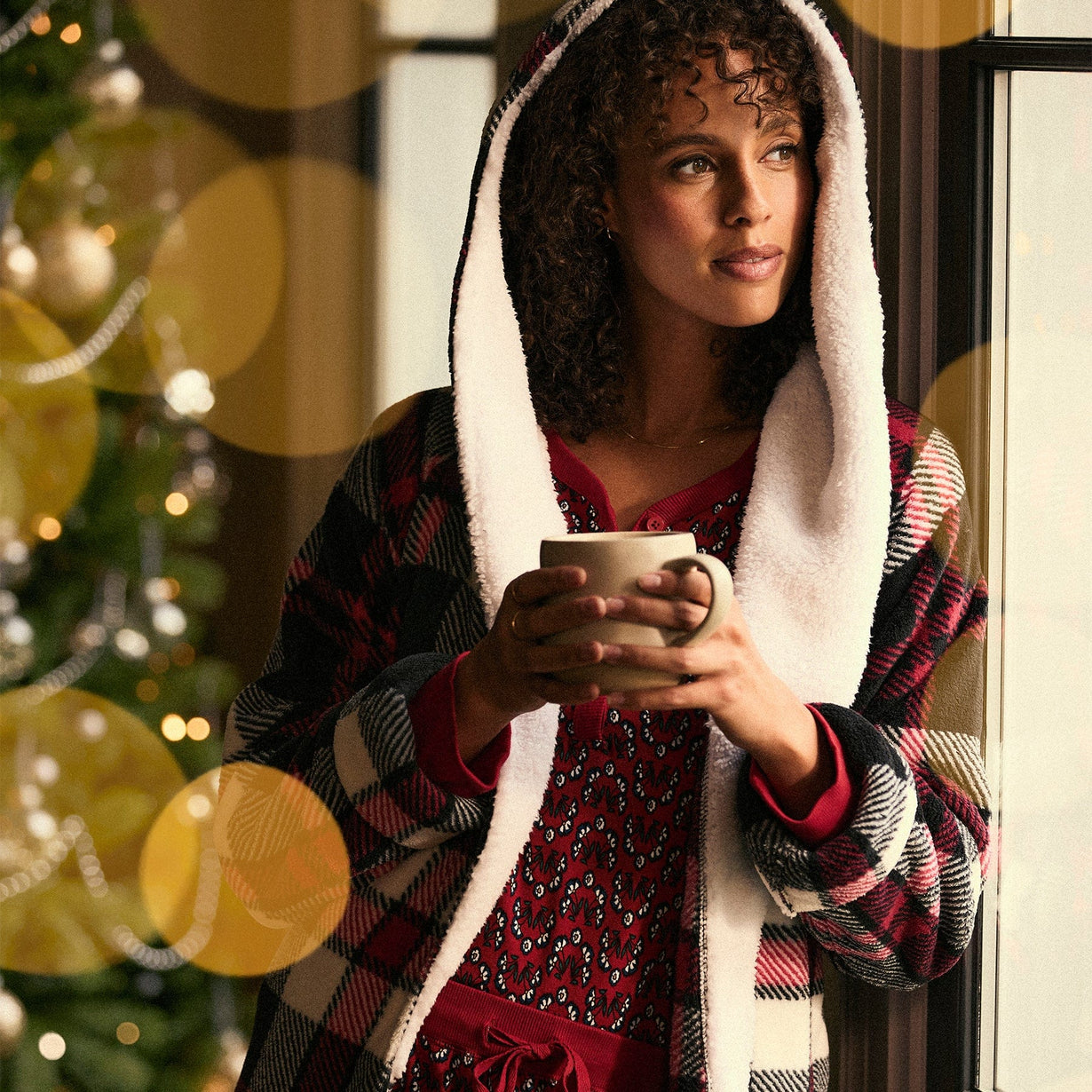 Woman standing next to window wearing red plaid fleece robe holding coffee mug