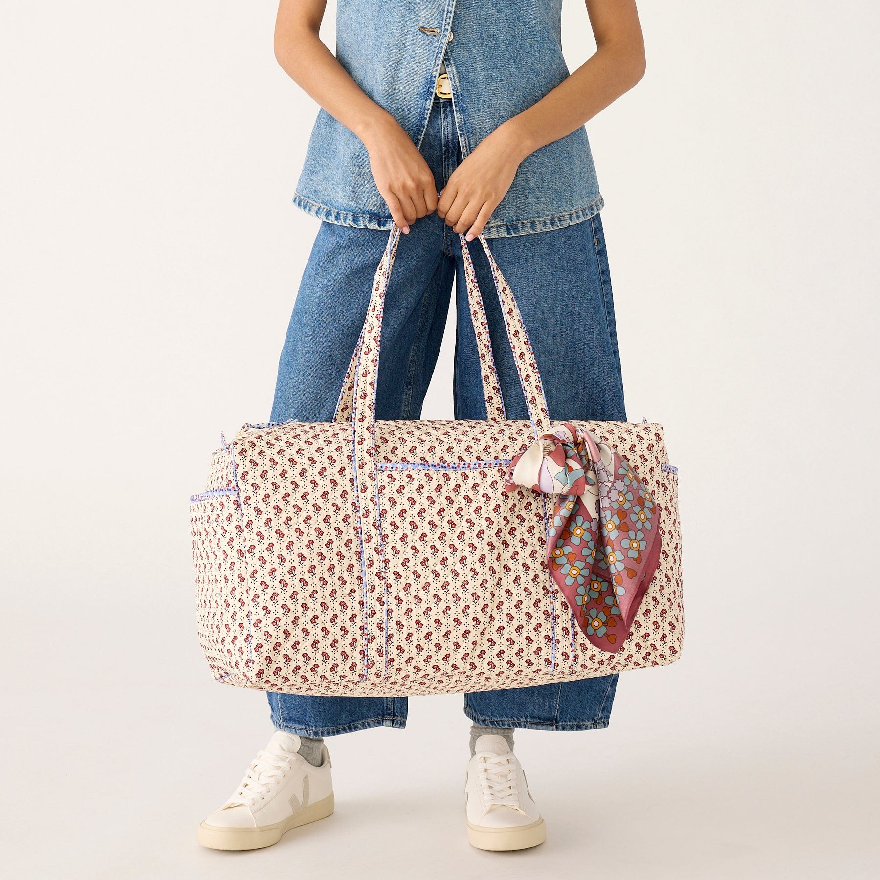 model wearing denim holding cream color large duffel bag with small red flowers pattern